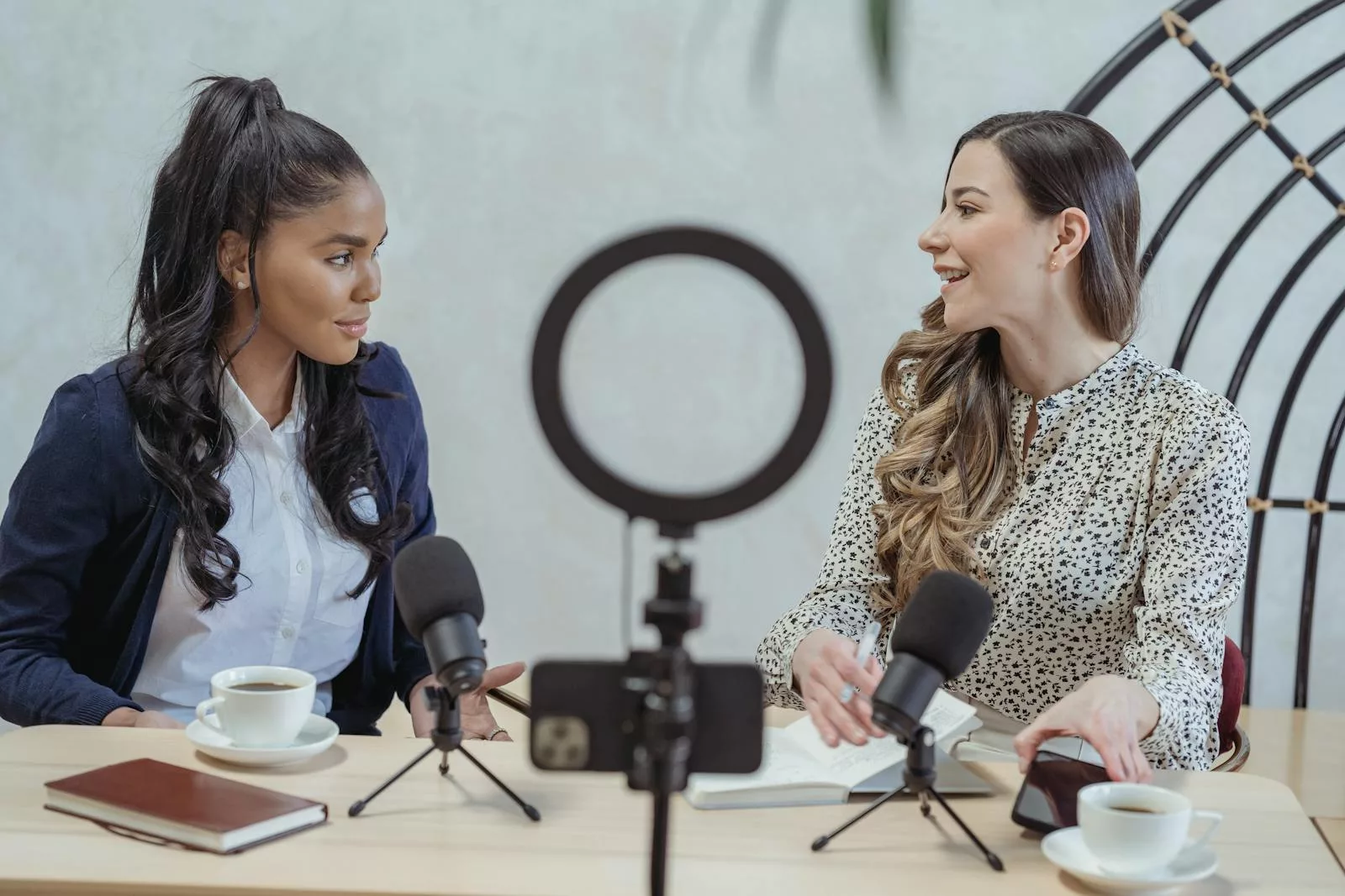 Positive blogger asking questions to African American speaker and recording interview on cellphone at table with microphones